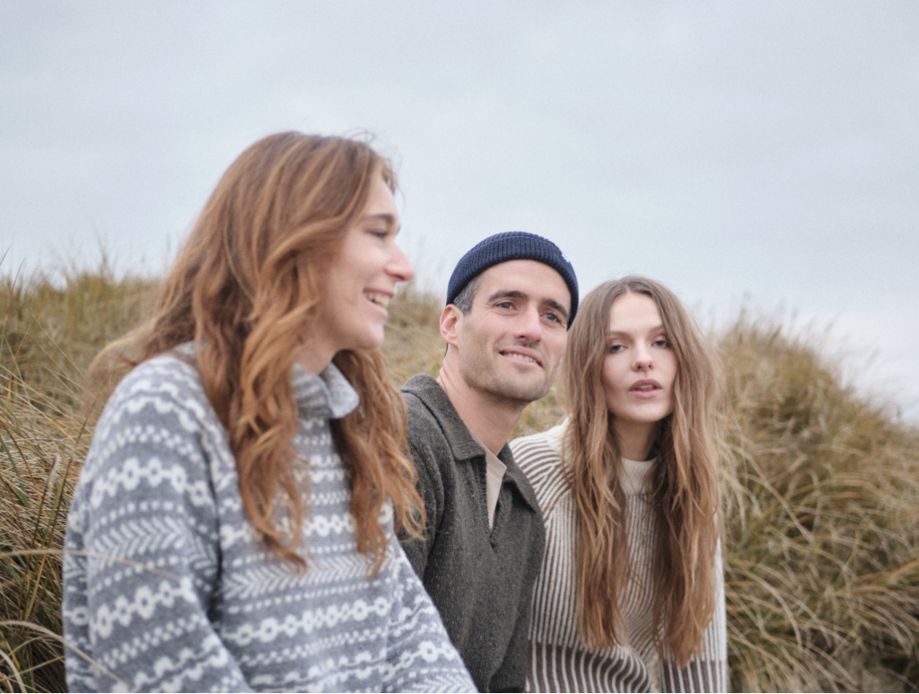 klitmoller-collective-skandinavische-mode 2 Frauen und ein Mann sitzen in einer Dünenlandschaft mit skandinavischen Wollstrickpuillovern von Klitmøller Collective