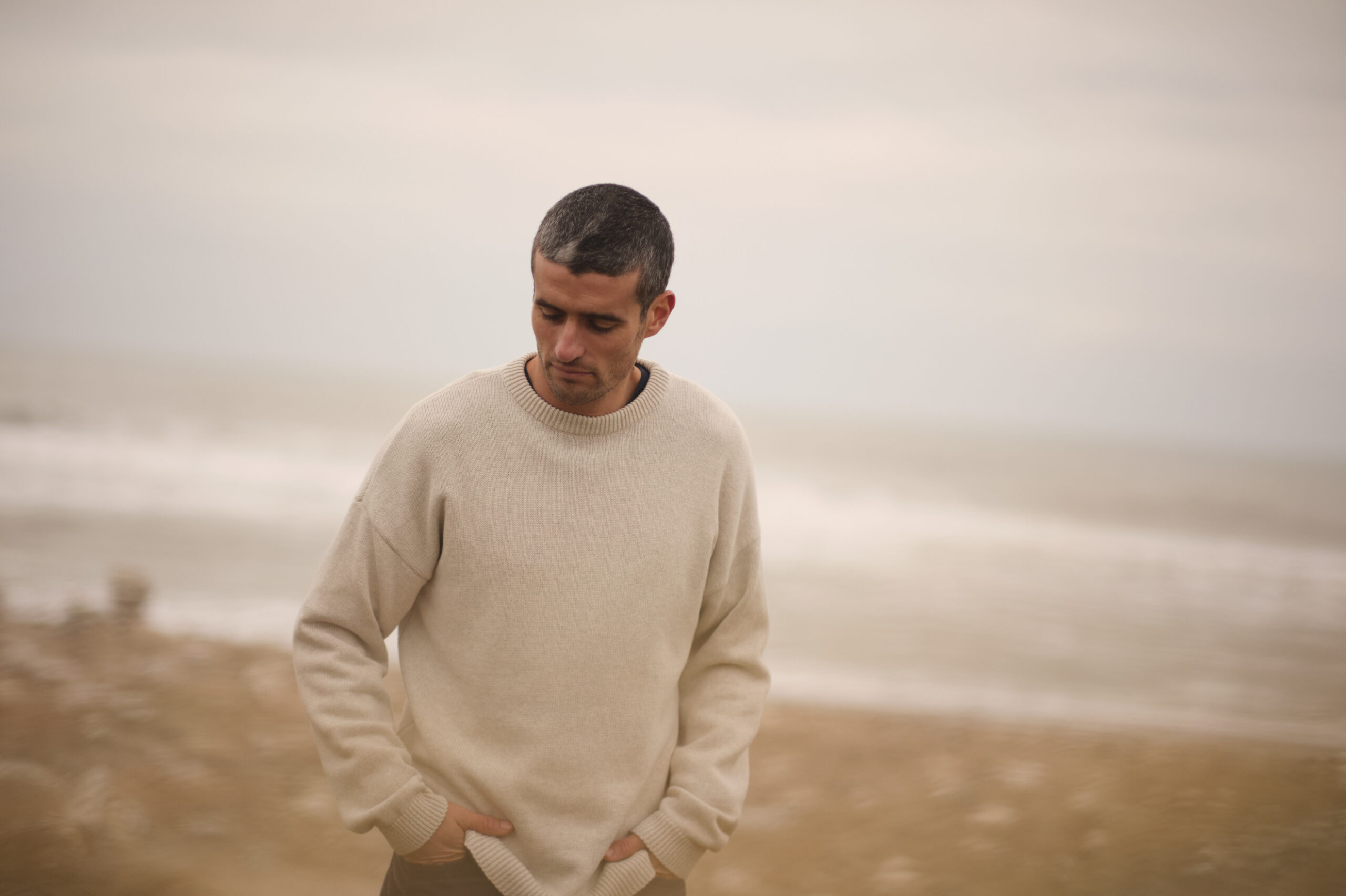 klitmoller-collective-zuendstoff-freiburg EIn Mann am Strand mit einem sandfarbenen Pullover des dänischen Labels Klitmøller Collective