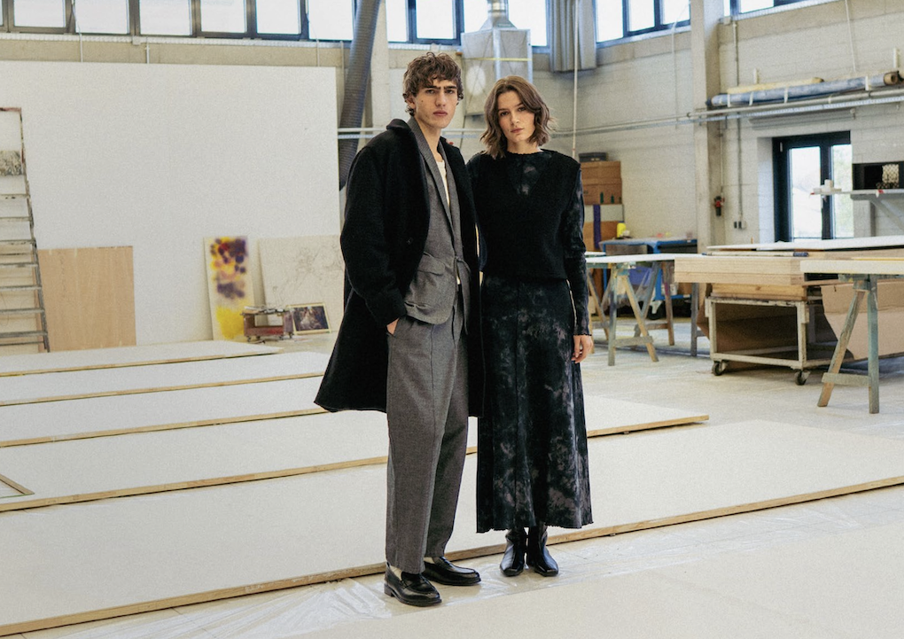 rotholz-mode-freiburg Frau und Mann mit Outfits von Rotholz stehen nebeneinander in einer Werkstatt und blicken in die Kamera